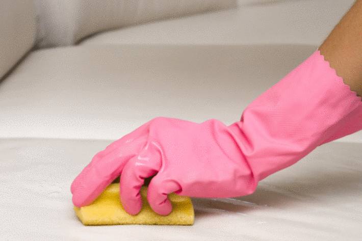 une main sous un gant en plastique rose enleve une tache avec une éponge sur un canapé blanc
