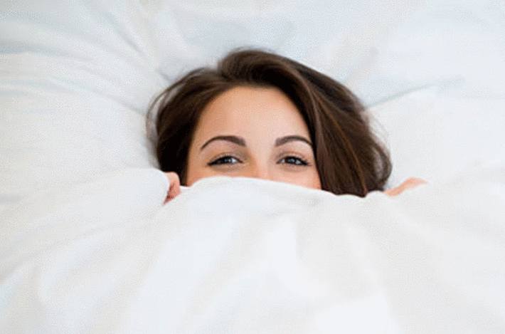 une femme brune dans son lit sous ses draps blancs