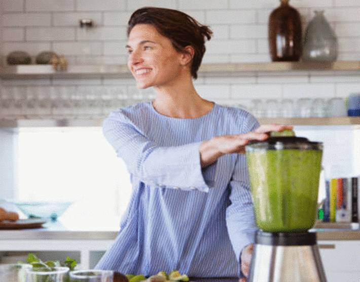 femme-brune-souriant-les-mains-sur-le-mixer-rempli-de-jus-de-légumes-verts
